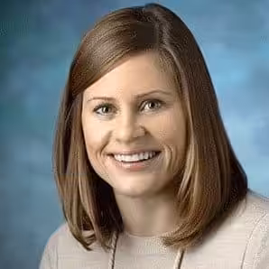 A person with shoulder-length brown hair and a beige top smiles warmly, possibly a professional associated with "Assessment and Treatment of Problem Behaviors within an Outpatient Clinic," posing against a blue background.