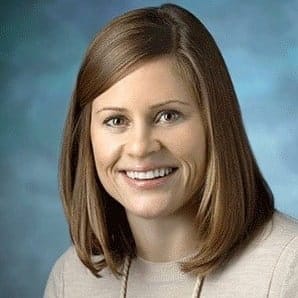 A person with shoulder-length brown hair and a beige top smiles warmly, possibly a professional associated with "Assessment and Treatment of Problem Behaviors within an Outpatient Clinic," posing against a blue background.