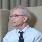 Man in glasses sitting indoors, wearing a tie.
