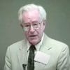 Elderly man wearing glasses and suit, speaking.