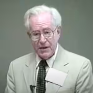 Elderly man wearing glasses and suit, speaking.