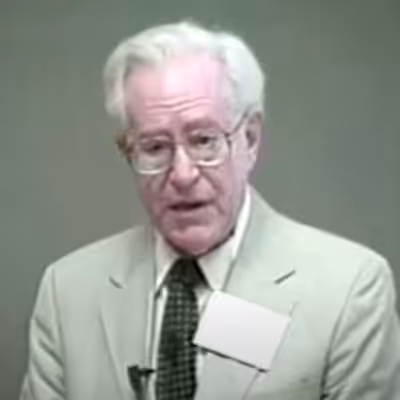 Elderly man wearing glasses and suit, speaking.