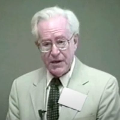 Elderly man wearing glasses and suit, speaking.
