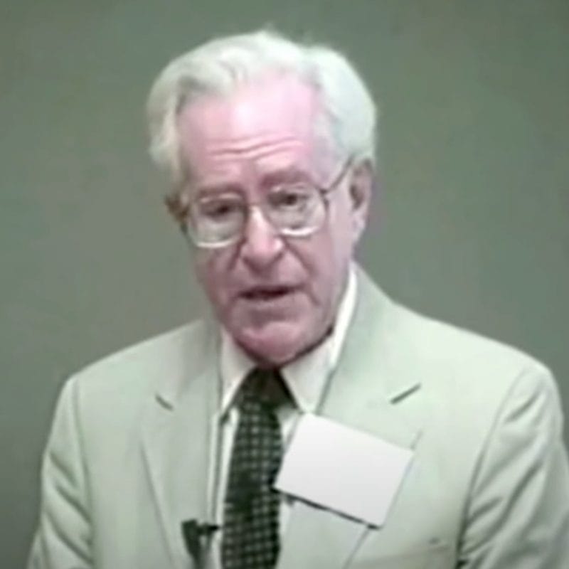 Elderly man wearing glasses and suit, speaking.