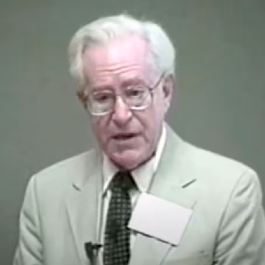 Elderly man wearing glasses and suit, speaking.